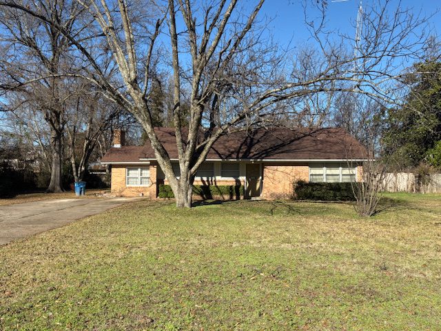 Single-story brick home with a brown roof, set on a spacious lawn. Leafless trees frame the house, and a blue trash can is visible by the driveway.